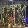 A woman stands confidently in front of a large sign displaying the letters "stk."