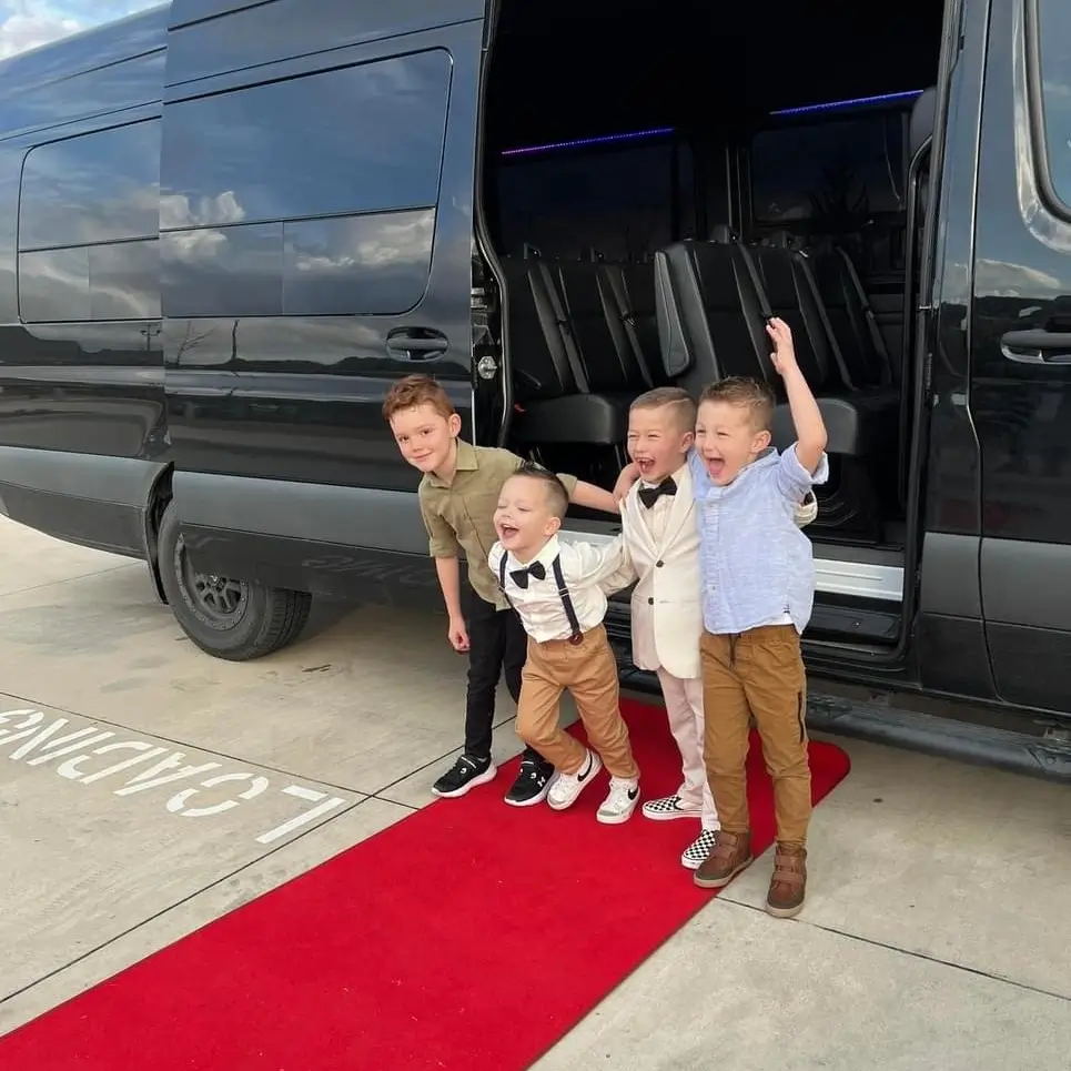 Three young boys pose on a red carpet, smiling in front of a sleek black limousine.