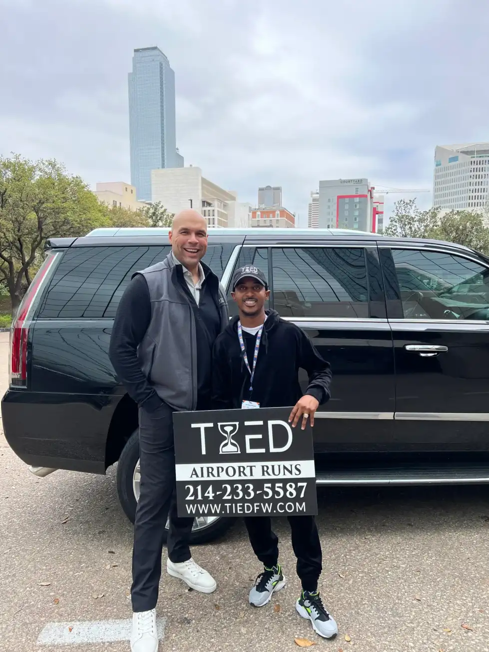 Two men stand beside a black SUV, which features a visible sign, in an outdoor setting.