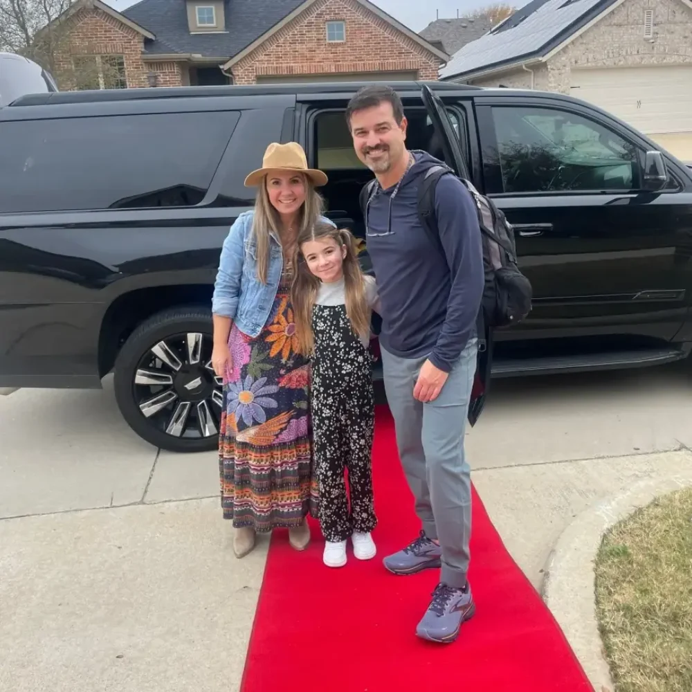 A man and woman pose on a red carpet beside a black SUV, showcasing elegance and style at a glamorous event.