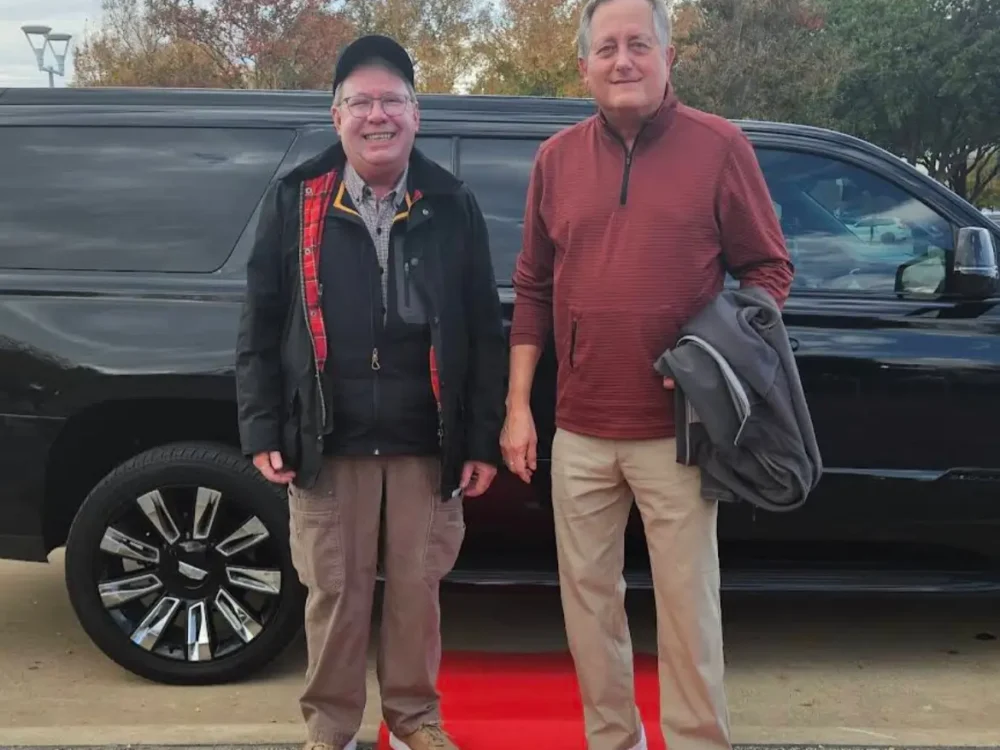 Two men pose on a red carpet beside a black SUV, showcasing a moment of elegance and style.